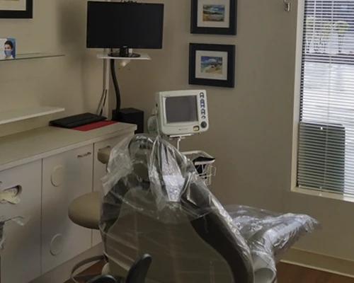Dental chair covered in protective plastic beside monitors, shelves, and window blinds in a clinical room.