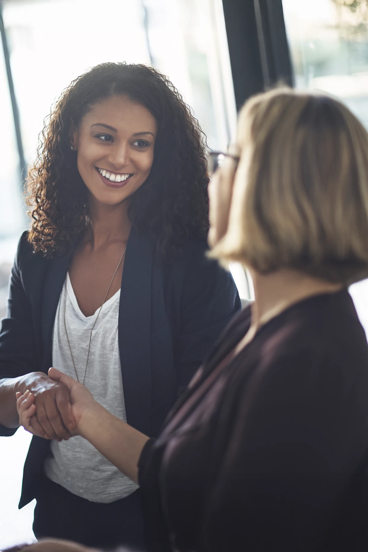 Two people shaking hands during a conversation inside an office setting with large windows in the background.